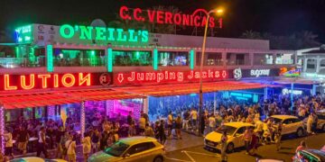 Escena de la calle nocturna con bares llenos de gente y autos estacionados.