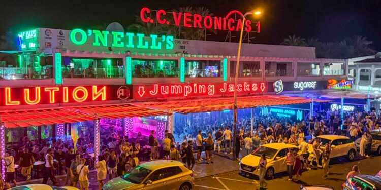 Escena de la calle nocturna con bares llenos de gente y autos estacionados.