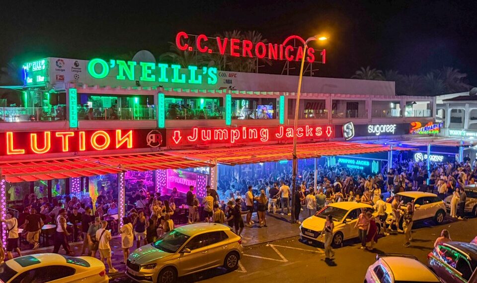 Escena de la calle nocturna con bares llenos de gente y autos estacionados.