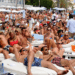 Escena de clubes de playa llenos de gente con gente descansando en camas de día blancas.