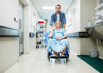 Mujer embarazada en una silla de ruedas siendo empujada por un hombre por un pasillo hospitalario.