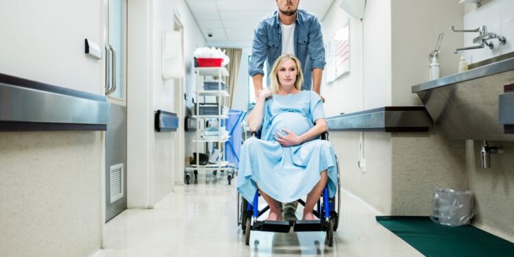 Mujer embarazada en una silla de ruedas siendo empujada por un hombre por un pasillo hospitalario.