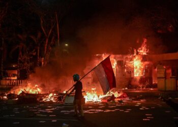 Un manifestante con una bandera indonesia durante una demostración donde un edificio está en llamas.