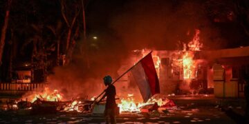Un manifestante con una bandera indonesia durante una demostración donde un edificio está en llamas.