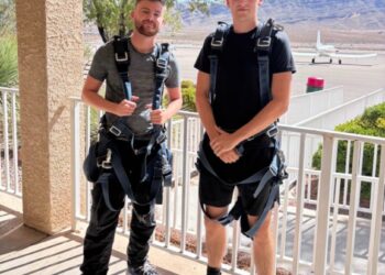 Dos hombres en arneses de paracaidismo posan para una foto con un pequeño avión y montañas en el fondo.