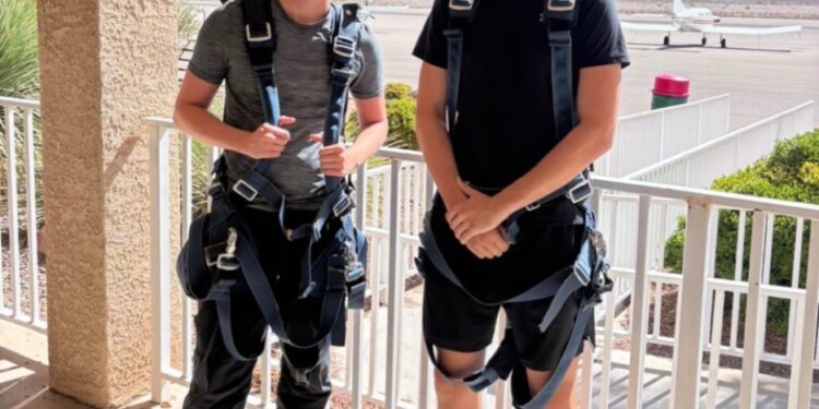 Dos hombres en arneses de paracaidismo posan para una foto con un pequeño avión y montañas en el fondo.