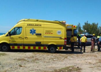 Ambulancia amarilla con sus puertas traseras abiertas en Cap Sant Pere Beach, con personal de emergencia reunido cerca.