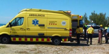 Ambulancia amarilla con sus puertas traseras abiertas en Cap Sant Pere Beach, con personal de emergencia reunido cerca.
