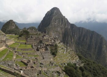 Sitio arqueológico de Machu Picchu con campos en terrazas y estructuras de piedra antiguas, debajo de una montaña cubierta de nubes.