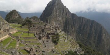 Sitio arqueológico de Machu Picchu con campos en terrazas y estructuras de piedra antiguas, debajo de una montaña cubierta de nubes.