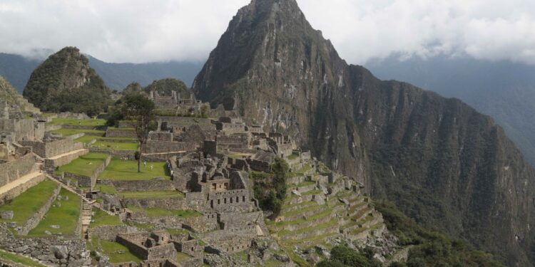 Sitio arqueológico de Machu Picchu con campos en terrazas y estructuras de piedra antiguas, debajo de una montaña cubierta de nubes.
