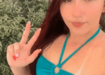 Una mujer con cabello largo y oscuro con un bikini azul e inferior, haciendo un signo de paz con su mano izquierda.