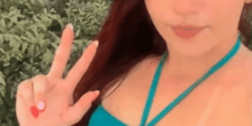 Una mujer con cabello largo y oscuro con un bikini azul e inferior, haciendo un signo de paz con su mano izquierda.