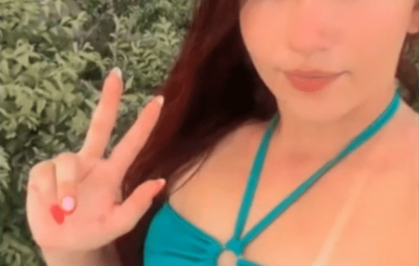 Una mujer con cabello largo y oscuro con un bikini azul e inferior, haciendo un signo de paz con su mano izquierda.