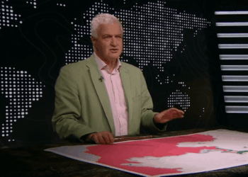 Un hombre con una chaqueta verde y gestos de camisa rosa mientras está de pie detrás de un escritorio con un mapa.