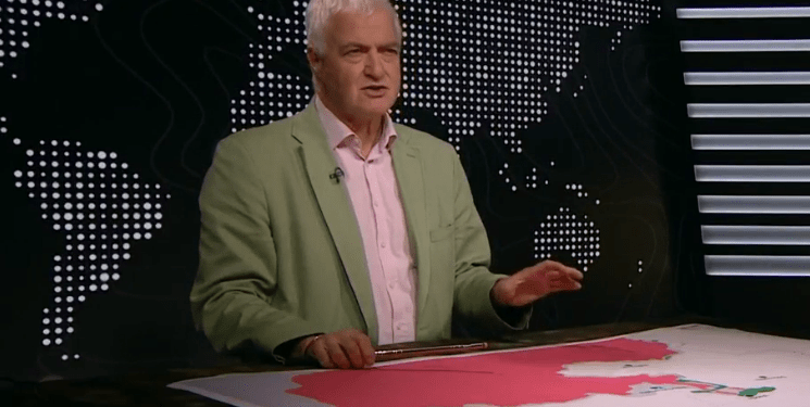 Un hombre con una chaqueta verde y gestos de camisa rosa mientras está de pie detrás de un escritorio con un mapa.