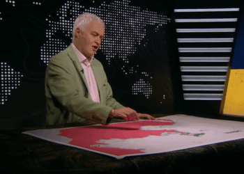 Un hombre con una chaqueta verde de pie sobre un mapa grande con un área rosa, señalando con las manos.