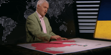 Un hombre con una chaqueta verde de pie sobre un mapa grande con un área rosa, señalando con las manos.