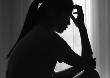 Silueta de una mujer joven sentada en una habitación oscura, cabeza abajo, sosteniendo su frente, que representa dolor o angustia.