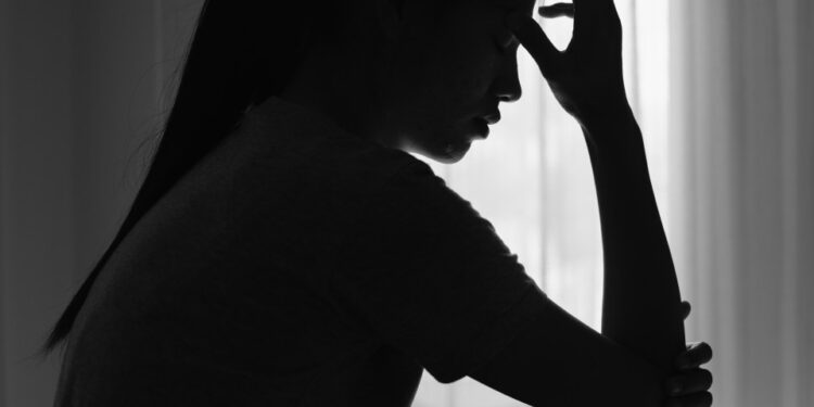 Silueta de una mujer joven sentada en una habitación oscura, cabeza abajo, sosteniendo su frente, que representa dolor o angustia.
