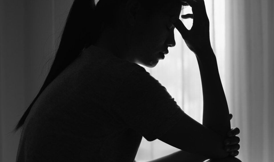 Silueta de una mujer joven sentada en una habitación oscura, cabeza abajo, sosteniendo su frente, que representa dolor o angustia.