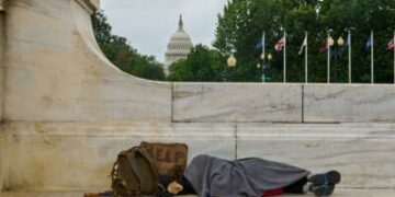 Una persona sin hogar duerme en el banco con el edificio del Capitolio de los Estados Unidos en el fondo.