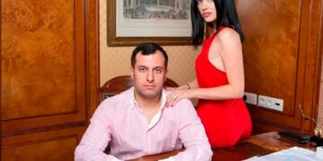 Boris Avakyan, de 43 años, con una camisa rayada, sentada en un escritorio de madera con su esposa Julia, con un vestido rojo, apoyado en su hombro.