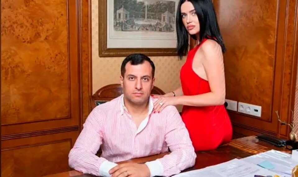 Boris Avakyan, de 43 años, con una camisa rayada, sentada en un escritorio de madera con su esposa Julia, con un vestido rojo, apoyado en su hombro.