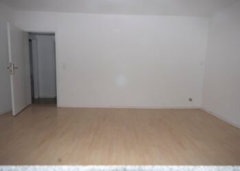 Una habitación vacía con paredes blancas, un piso de madera claro y una puerta abierta que conduce a otra habitación.