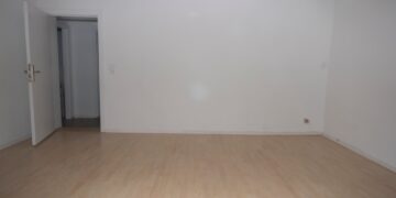 Una habitación vacía con paredes blancas, un piso de madera claro y una puerta abierta que conduce a otra habitación.