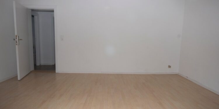 Una habitación vacía con paredes blancas, un piso de madera claro y una puerta abierta que conduce a otra habitación.