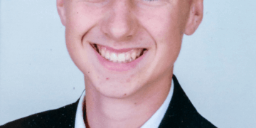 Un joven sonriente con un traje negro y una corbata roja con una camisa blanca.