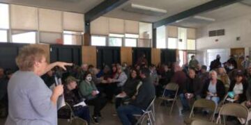 Después del incendio de Dixie de 2021, el colectivo de Indian Valley se reúne para su reunión mensual en la cafetería de la Escuela Primaria Greenville, uno de los pocos edificios en Greenville, California, para sobrevivir. Estas reuniones son donde los miembros de la comunidad comparten información vital sobre los esfuerzos de recuperación y los nuevos proyectos. Sue Weber (quien escribió una pieza en advertencia de la bandera roja) está hablando.