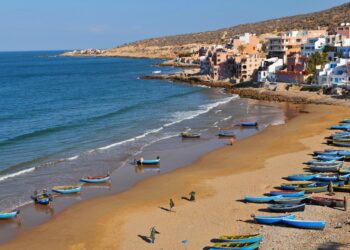 Vista aérea de Taghazout, Marruecos, con un pueblo construido en una ladera con vistas a una playa salpicada de barcos de pesca azul y amarillo.