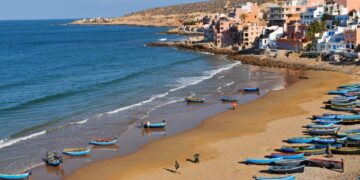 Vista aérea de Taghazout, Marruecos, con un pueblo construido en una ladera con vistas a una playa salpicada de barcos de pesca azul y amarillo.