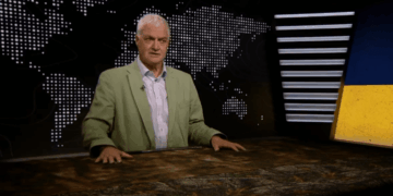 El hombre con chaqueta verde se sienta en un escritorio que da una presentación frente a un mapa mundial.