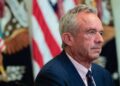 Robert F. Kennedy Jr. escucha durante un evento sobre tecnología de salud en la sala este de la Casa Blanca con una bandera estadounidense detrás de él.