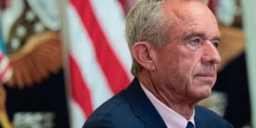 Robert F. Kennedy Jr. escucha durante un evento sobre tecnología de salud en la sala este de la Casa Blanca con una bandera estadounidense detrás de él.