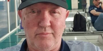 John Murphy, un veterano británico, con una gorra negra y una camisa a rayas, sentada en lo que parece ser un aeropuerto o sala de espera pública.