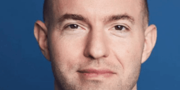 Tiro en la cabeza de un hombre con cabeza afeitada y barba ligera, con traje y sonriendo.