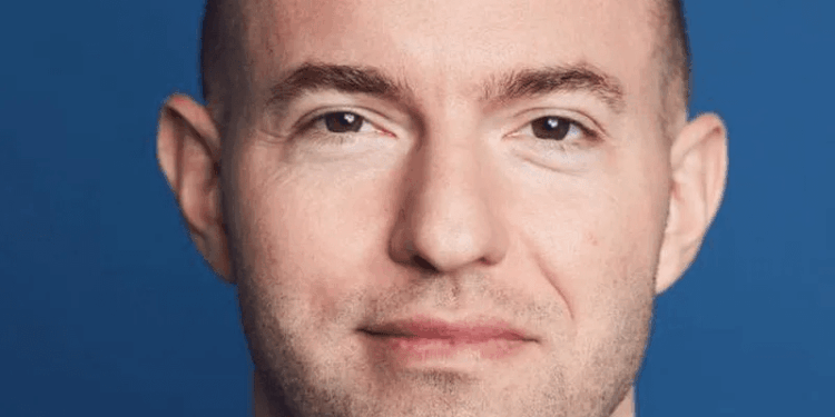 Tiro en la cabeza de un hombre con cabeza afeitada y barba ligera, con traje y sonriendo.