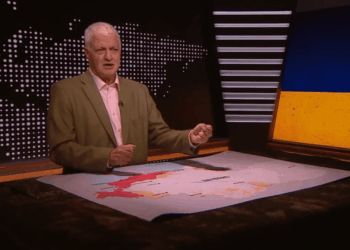 Un hombre que informa sobre las noticias con un mapa de Ucrania en una mesa frente a él, y una imagen de la bandera ucraniana detrás de él.