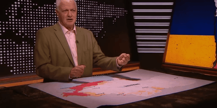 Un hombre que informa sobre las noticias con un mapa de Ucrania en una mesa frente a él, y una imagen de la bandera ucraniana detrás de él.