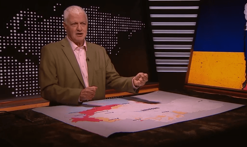 Un hombre que informa sobre las noticias con un mapa de Ucrania en una mesa frente a él, y una imagen de la bandera ucraniana detrás de él.
