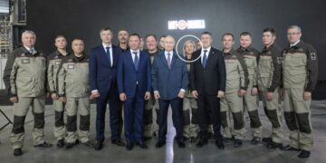 Una mujer rubia, rodeada de blanco, se encuentra entre los trabajadores de la fábrica y los funcionarios, incluido Vladimir Putin, en las plantas de defensa de Motovilikha.