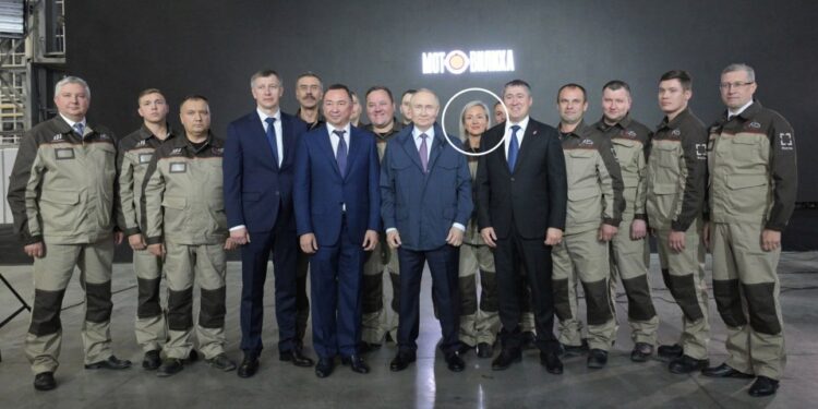 Una mujer rubia, rodeada de blanco, se encuentra entre los trabajadores de la fábrica y los funcionarios, incluido Vladimir Putin, en las plantas de defensa de Motovilikha.