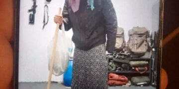 Un hombre con ropa de mujer, que incluye un pañuelo rosado y una falda estampada, sosteniendo un palo y una bolsa de plástico blanca, de pie en una habitación con equipo militar.
