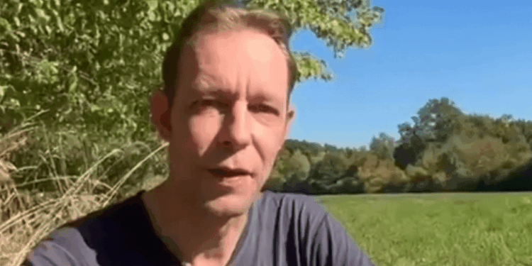Un hombre que usa una camiseta oscura que habla con la cámara al aire libre con follaje verde y cielo azul en el fondo.