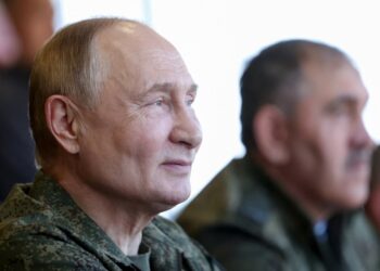 El presidente ruso, Vladimir Putin, con un uniforme militar, mirando hacia la derecha, con otro hombre borroso en el fondo.