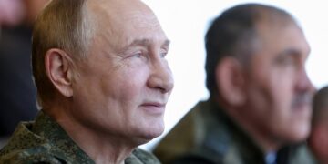 El presidente ruso, Vladimir Putin, con un uniforme militar, mirando hacia la derecha, con otro hombre borroso en el fondo.